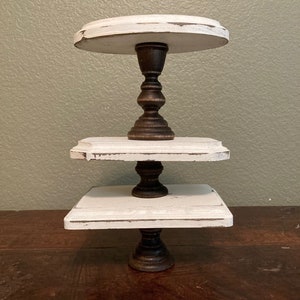 May include: A three-tiered, distressed white wooden cake stand with dark brown, ornate pedestals. The top tier is round, the middle tier is rectangular, and the bottom tier is square. The stand is on a dark brown wooden surface.
