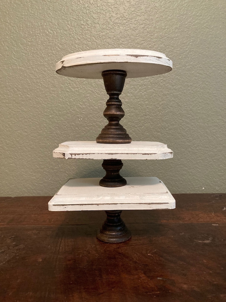 May include: A three-tiered, distressed white wooden cake stand with dark brown, ornate pedestals. The top tier is round, the middle tier is rectangular, and the bottom tier is square. The stand is on a dark brown wooden surface.