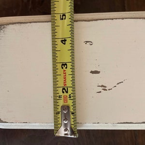 May include: A rectangular, off-white wooden sign with distressed edges. The sign is approximately 10 cm wide, as measured by a yellow tape measure. The sign has a slightly worn appearance, with some paint chipping.