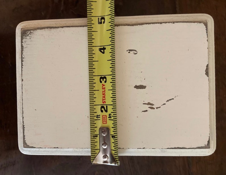 May include: A rectangular, off-white wooden sign with distressed edges. The sign is approximately 10 cm wide, as measured by a yellow tape measure. The sign has a slightly worn appearance, with some paint chipping.