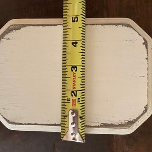 May include: A rectangular, off-white wooden sign with distressed edges, measuring approximately 12.7 cm in length. A yellow tape measure is positioned vertically, providing a size reference. The sign has a rustic, aged appearance.