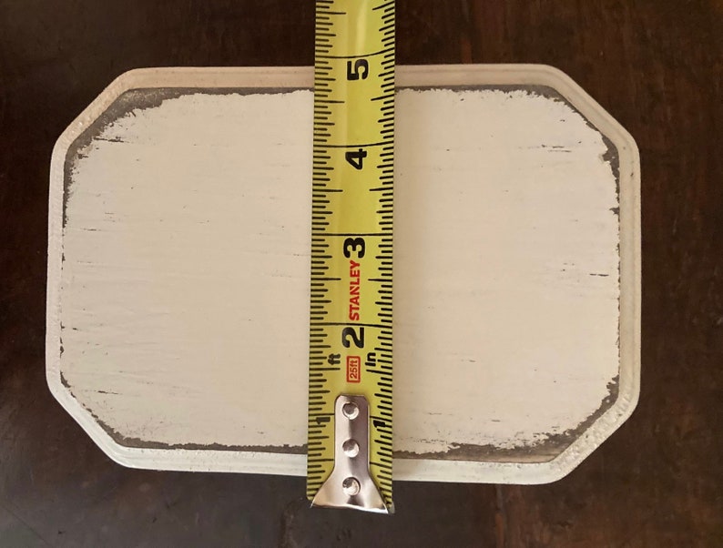 May include: A rectangular, off-white wooden sign with distressed edges, measuring approximately 12.7 cm in length. A yellow tape measure is positioned vertically, providing a size reference. The sign has a rustic, aged appearance.