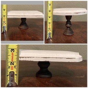 May include: A small, distressed white and black cake stand. The stand has a round top and a dark, turned pedestal base. The height is approximately 10 cm.