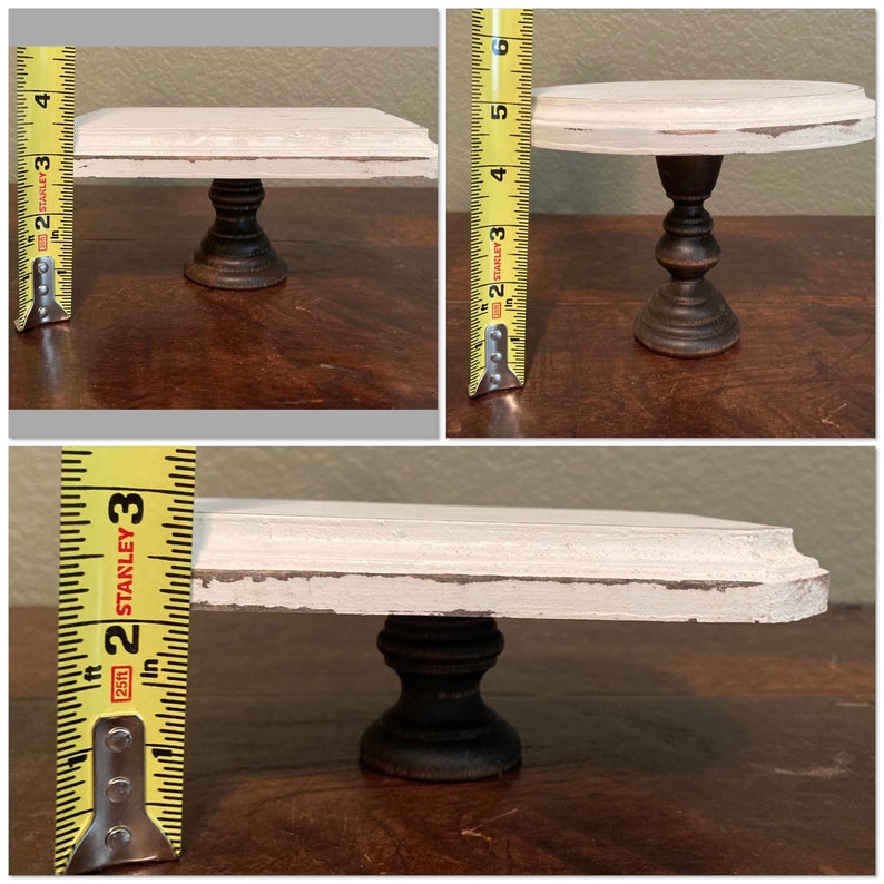 May include: A small, distressed white and black cake stand. The stand has a round top and a dark, turned pedestal base. The height is approximately 10 cm.