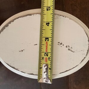 May include: An oval, distressed white wooden tray with a grey trim. A yellow tape measure is placed vertically on the tray, showing a measurement of approximately 12.7 cm. The tray has a rustic, vintage aesthetic.