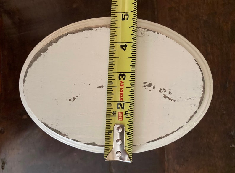 May include: An oval, distressed white wooden tray with a grey trim. A yellow tape measure is placed vertically on the tray, showing a measurement of approximately 12.7 cm. The tray has a rustic, vintage aesthetic.