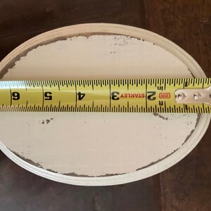 May include: An oval, off-white painted wooden tray with distressed edges. A yellow tape measure shows the tray is approximately 15 cm long. The tray is on a dark brown surface.