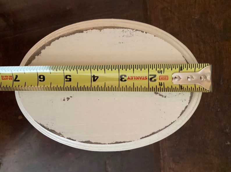 May include: An oval, off-white painted wooden tray with distressed edges. A yellow tape measure shows the tray is approximately 15 cm long. The tray is on a dark brown surface.