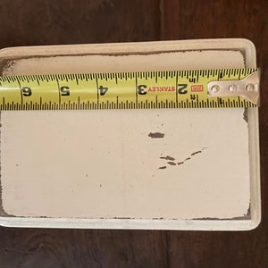May include: A rectangular, cream-coloured painted wooden sign with distressed edges. The sign is approximately 15 cm long, as measured by a yellow Stanley tape measure. The sign is on a dark brown wooden surface.