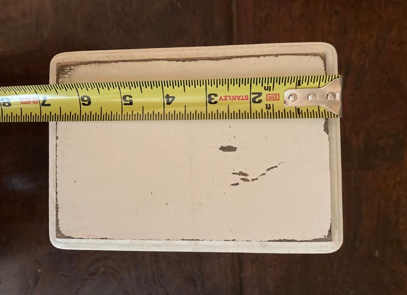May include: A rectangular, cream-coloured painted wooden sign with distressed edges. The sign is approximately 15 cm long, as measured by a yellow Stanley tape measure. The sign is on a dark brown wooden surface.