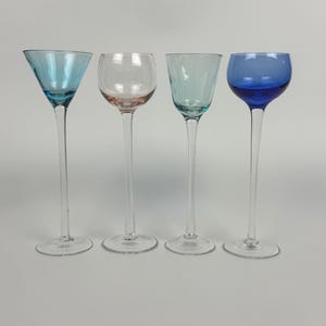 May include: Four colorful stemmed glasses. The glasses have long, slender stems and varying bowl shapes. The bowls are in shades of blue, pink, and clear. The glasses are arranged in a row against a plain background.