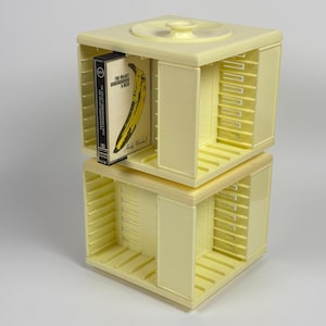May include: A two-tiered, pale yellow cassette tape storage unit. The unit has a square shape with open compartments and a central handle on top. A cassette tape with a banana graphic is visible in one compartment. The text on the cassette reads "The Velvet Underground & Nico."