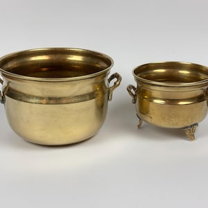 May include: Two polished brass bowls with rounded bodies and small side handles. The larger bowl has a smooth base, while the smaller bowl features three decorative feet. Both containers have a metallic gold finish and a wide, open top design.