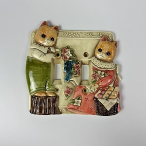 May include: A ceramic double light switch plate featuring two anthropomorphic cats in relief. One cat wears green overalls and stands on a brown stump, while the other wears a red dress and sits by a basket. Floral accents and a blue ribbon are present.