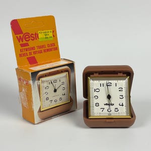 May include: A brown Westclox keywound travel clock with a square face, black numerals, and gold-tone trim, displayed open next to its original orange and white cardboard packaging featuring the text Westclox Keywound Travel Clock.