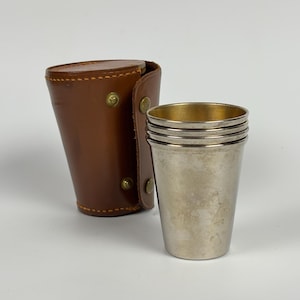 May include: Four silver metal cups with gold interiors, beside a brown leather case. The case is cylindrical with brass rivets and a snap closure. The cups are stacked. The case is made of brown leather.
