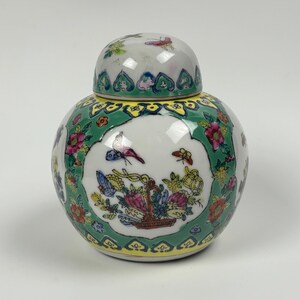 May include: A round, white ceramic jar with a lid, decorated with colourful floral and butterfly designs. The jar has a green border with yellow accents and a detailed illustration of a basket of flowers. The lid mirrors the jar's design.