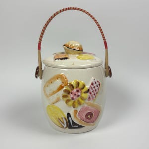 May include: Cream-colored ceramic cookie jar with a lid and a woven handle. The jar is decorated with colorful, raised relief images of baked goods, including bread, flowers, and pastries. The handle is made of woven wood with red accents.