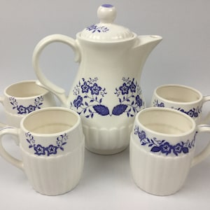 May include: A white ceramic teapot with a blue floral design and four matching mugs. The teapot has a lid with a knob on top. The mugs have handles and a blue floral design around the rim.