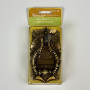 May include: A brass-colored door knocker in a clear plastic package. The knocker has an ornate design with a central bar and a handle. The packaging includes the text "Door Knocker" and "Marteau de Porte".