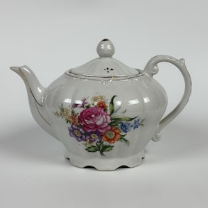 Peut inclure: Théière en porcelaine blanche avec un motif floral composé de fleurs roses, violettes et oranges. La théière a une anse incurvée, un bec verseur et un couvercle avec un petit bouton. Des bordures dorées ornent le bec et le couvercle.