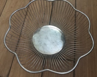 May include: A silver-colored, wire fruit basket with a scalloped edge. The basket has a circular, solid base and radiating wires that form the sides. The basket is empty and sits on a wooden surface.