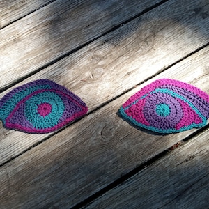 Puede incluir: Dos ojos tejidos a crochet con hilo rosa, morado y turquesa. Los ojos están sobre una superficie de madera.