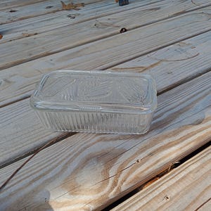 Puede incluir: Una fuente rectangular de vidrio transparente con tapa. La fuente tiene un diseño acanalado en los lados y un patrón decorativo en la tapa. La fuente está sobre una superficie de madera.