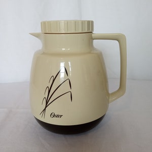 Puede incluir: Una jarra térmica beige y marrón con tapa blanca y un diseño estilizado de espigas de trigo. La jarra está etiquetada con "Oster".