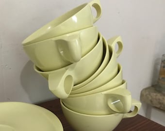 May include: A stack of six pale yellow ceramic teacups with handles. The cups are stacked in a staggered pattern, with the top cup tilted slightly to the side. The cups are all the same size and shape, and they have a smooth, glossy finish.