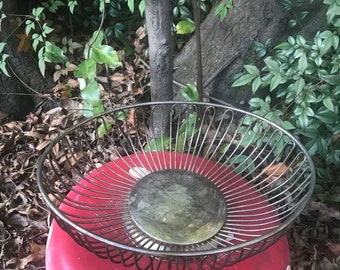May include: A silver wire basket with a round base sits on a red and white mushroom-shaped object. The basket is made of thin wire and has a circular design.