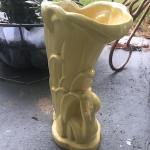 Pode incluir: Um vaso de cerâmica amarelo pálido com uma parte superior alargada e um design orgânico esculpido. O vaso tem um acabamento liso e brilhante e uma base arredondada. O vaso está sobre uma superfície cinzenta.
