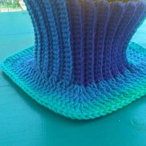 May include: A crocheted neck warmer with a gradient of blue and green colors. The neck warmer is made with a textured stitch and has a wide, folded brim.