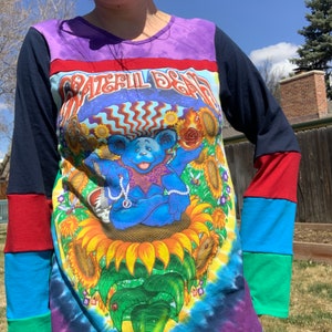 May include: A tie-dye long-sleeve shirt with a Grateful Dead graphic featuring a blue bear sitting on a sunflower. The shirt has a multi-colored patchwork design on the sleeves.