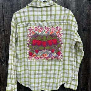 May include: Green and white plaid button-down shirt with a strawberry patch on the back. The patch features red strawberries with green leaves and floral accents. The shirt is hanging on a white hanger against a wooden fence.