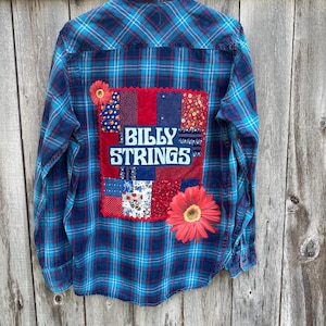 May include: Blue and red plaid flannel shirt with a patchwork design on the back. The patchwork includes a red fabric square with the text "BILLY STRINGS" in white letters. There are also floral fabric squares and a large red flower.