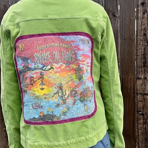 May include: A green denim jacket with a colorful patch on the back. The patch features the text "Widespread Panic Snake Oil King" with an illustration of cartoon characters and a vibrant sunset scene. The jacket has a button-down front and long sleeves.
