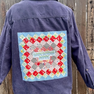 May include: A dark blue denim jacket with a colorful patchwork design on the back. The patchwork features a square pattern with red, white, and black squares, and the words "WIDESPREAD PANIC" are stitched in the center.