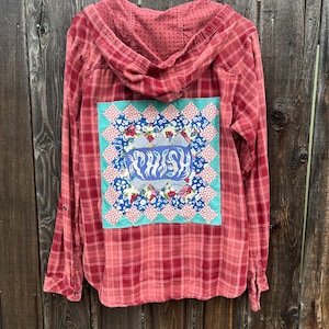May include: Red plaid button-down shirt with a patchwork design on the back. The patchwork features a blue and floral pattern with the word "Wish" in the center. The shirt has a hood and is hanging on a white hanger.