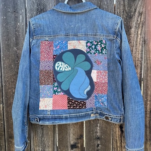 May include: A blue denim jacket with a patchwork design on the back. The patchwork includes a variety of floral patterns and a large green and blue patch with the word "PHISH" in white letters.