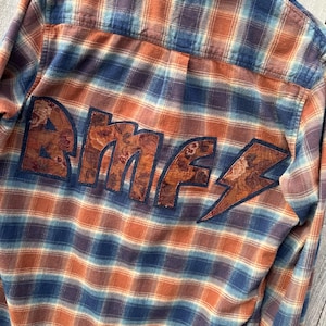 May include: A blue and orange plaid flannel shirt with a floral patterned patch that reads "BMF" on the back.