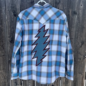 May include: A blue and white plaid flannel shirt with a large, dark blue and burgundy lightning bolt patch on the back.