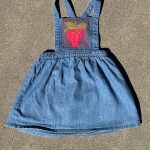 May include: A blue denim overall dress featuring a square bib with a rectangular patch. The patch displays a graphic of a red strawberry with green leaves. The dress has a gathered skirt and two shoulder straps that cross at the back.