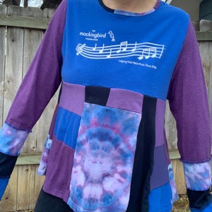 May include: A long-sleeved shirt with a patchwork design in shades of purple, blue, and black. The shirt features a white graphic of a bird and musical notes with the text "The Mockingbird Foundation" and "Helping Kids Make Music Since 1996".