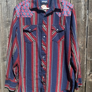 May include: A long-sleeved, striped button-down shirt with a red and blue color scheme. The shirt has a Wrangler label on the chest and a unique donut pattern on the shoulders.