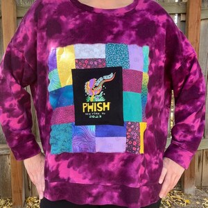 May include: Purple tie-dye sweatshirt with a patchwork design featuring a Phish logo patch. The logo reads "Phish" with the text "New York, NY 2023" underneath.