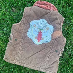 May include: A brown corduroy vest with a quilted design. The vest has a white crocheted butterfly applique with a red lightning bolt design in the center.