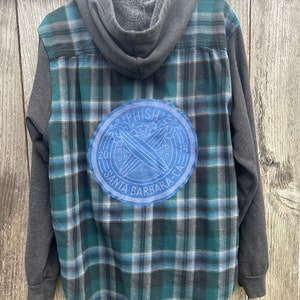 May include: A hooded flannel shirt with a blue, green, and gray plaid pattern. The sleeves and hood are gray. A blue circular patch on the back features the text "PHISH" and "SANTA BARBARA CA".