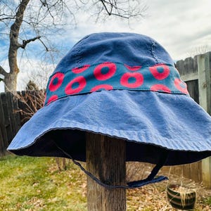 May include: A blue bucket hat with a wide brim and a chin strap. The hat features a band with a teal background and a pattern of red donut shapes. The hat is made of a soft, durable fabric and is suitable for outdoor wear.