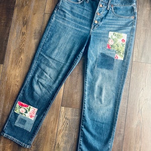 May include: Blue denim jeans with floral patches on the knees. The jeans have a button fly and a classic five-pocket design. The patches feature a colorful floral print with red and pink flowers.